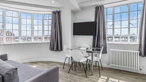 A modern living room with a grey sofa, a glass dining table with white chairs, a flat-screen TV mounted on the wall, and large windows with grey curtains. - Chelsea Cloisters Serviced Apartments (London)