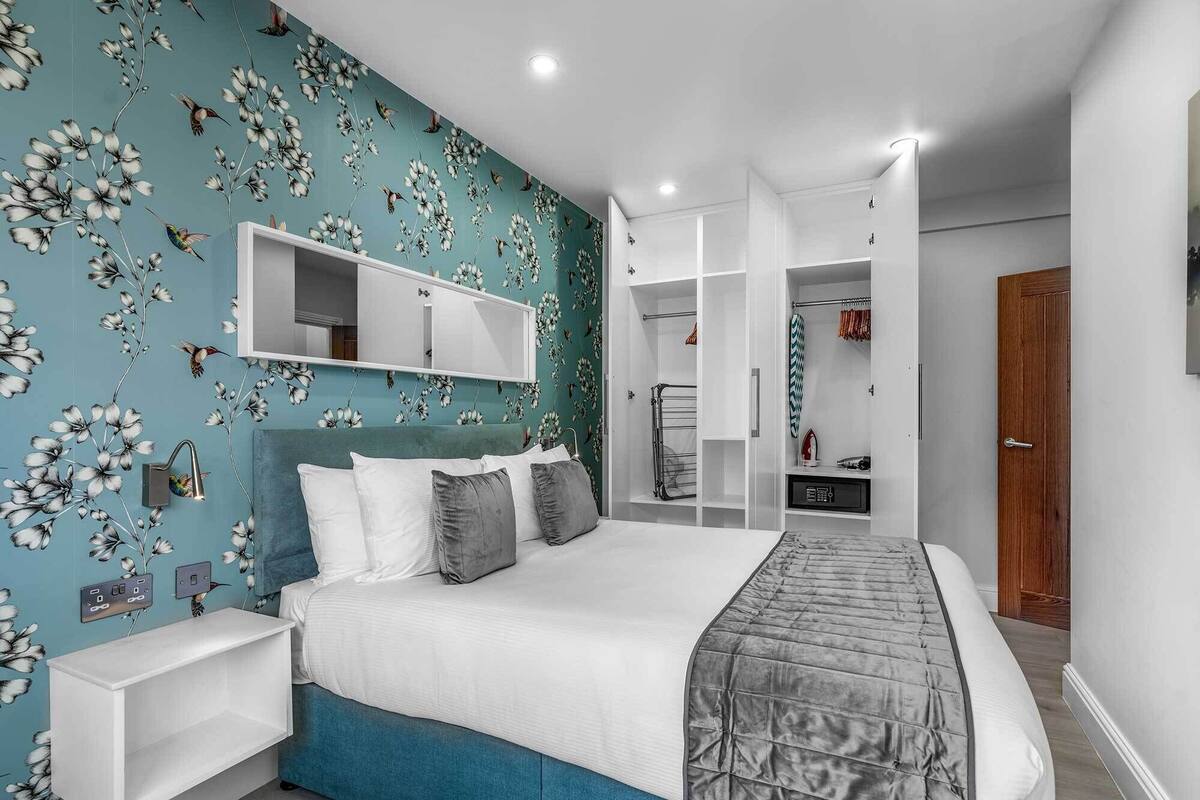 Une chambre moderne avec un papier peint à motifs floraux de couleur turquoise, un grand lit avec une literie grise, une table de chevet blanche et des étagères intégrées.