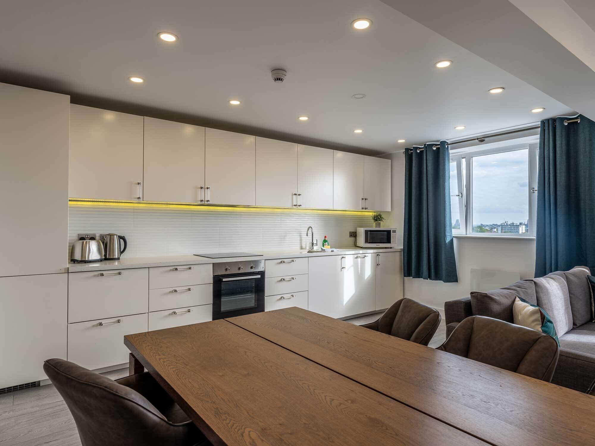 A modern kitchen with white cabinets, a wooden dining table, and a view of the city through a window.