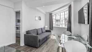 A modern living room with a grey sofa, a glass dining table with white chairs, and a large window with curtains. - Chelsea Cloisters Serviced Apartments (London)