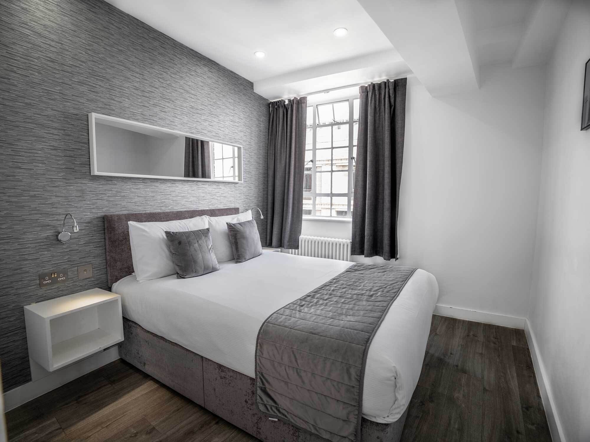 A modern bedroom with a large bed, a built-in nightstand, a wall-mounted mirror, and a window with curtains.