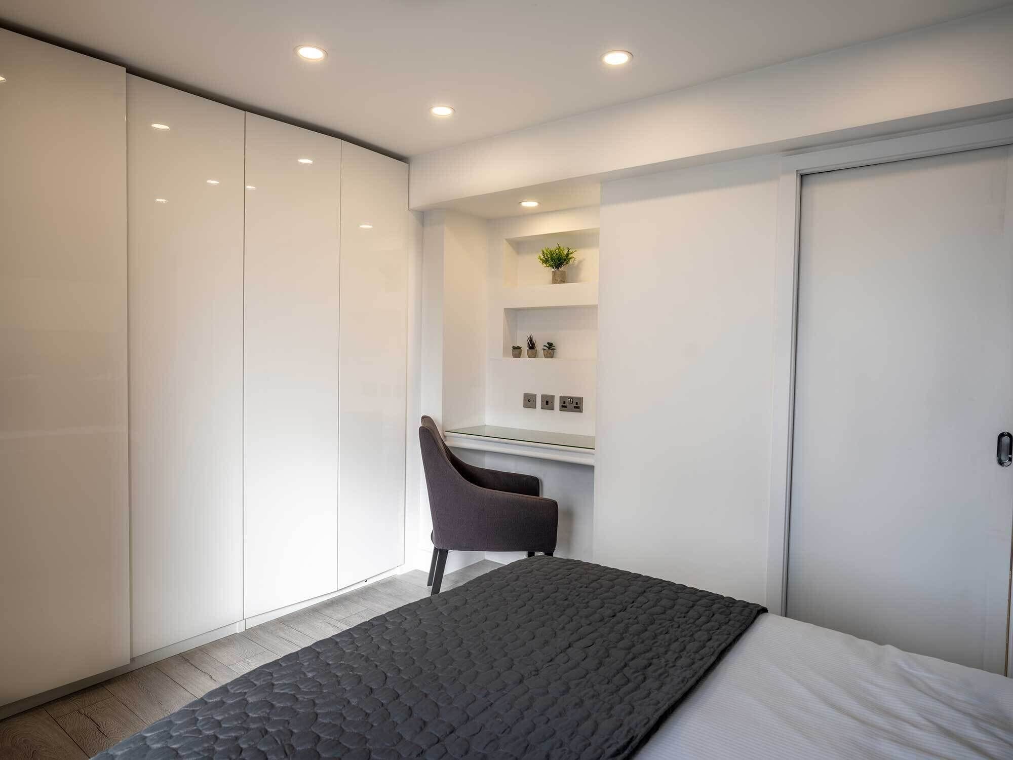 Un dormitorio con cama, escritorio y silla. Se ve un armario y una estantería con plantas.