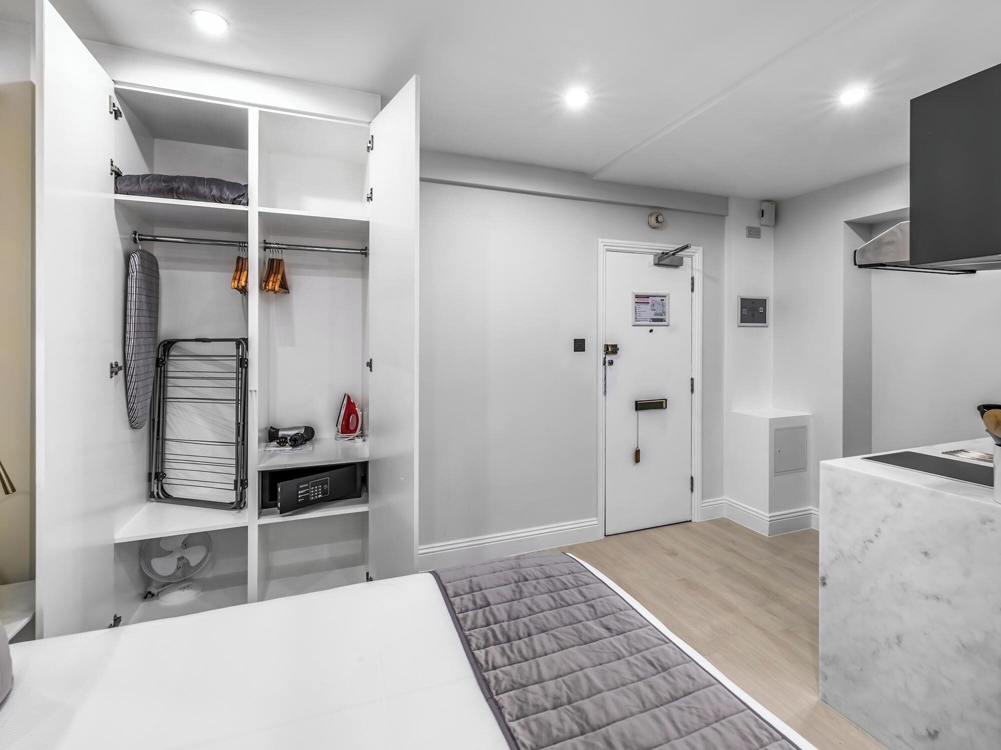 Camera da letto moderna con un letto, un armadio pieno di vestiti, una piccola tavola da stiro e una porta con serratura.