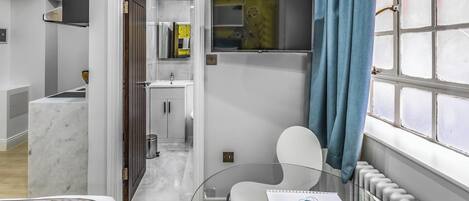 Habitación de hotel moderna con cama, una mesita de vidrio con un control remoto, una silla, televisor montado en la pared y vista a la ciudad a través de una ventana.