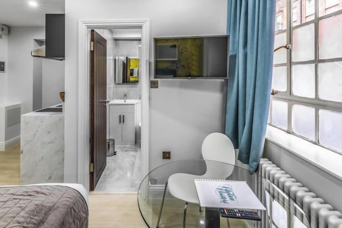 Una habitación de hotel moderna con una cama, una mesita de vidrio con un cuaderno, una silla, un televisor montado en la pared y vistas de la ciudad a través de amplias ventanas.