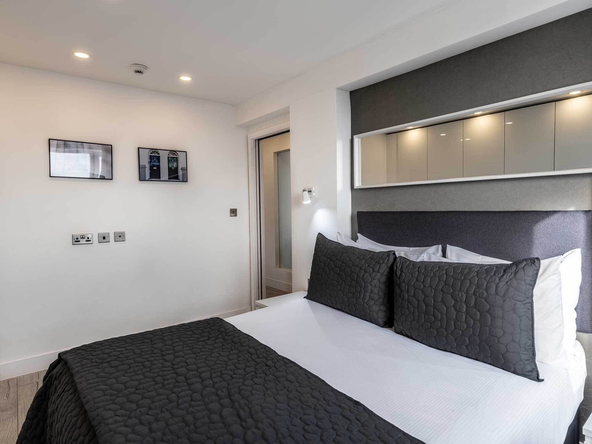 A modern hotel room with a large bed, a dark headboard, and a large mirror above the bed. There are two wall-mounted light switches and a door to the right.