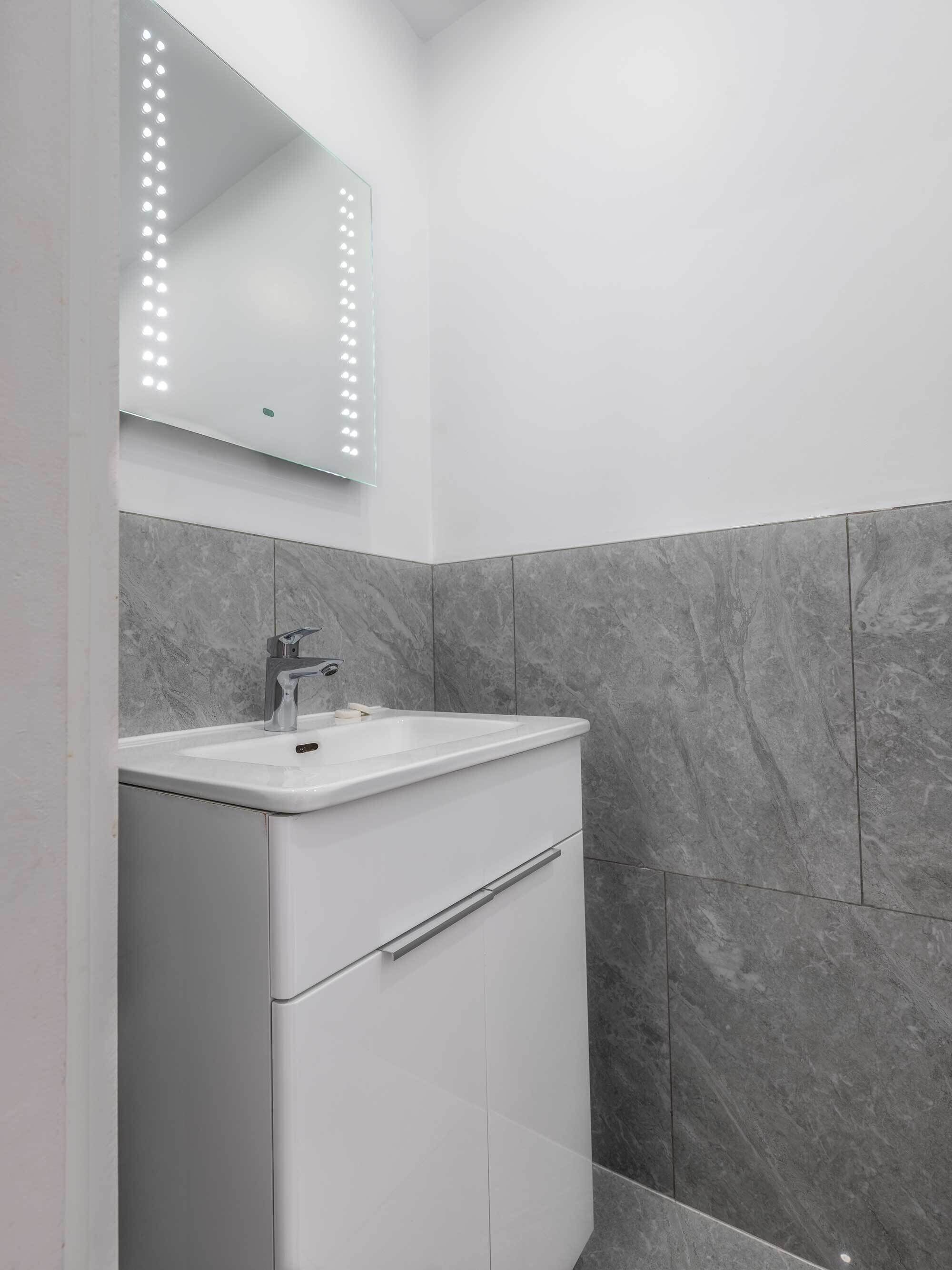 A modern bathroom with a white vanity, a mirror with lights, and grey marble tiles.