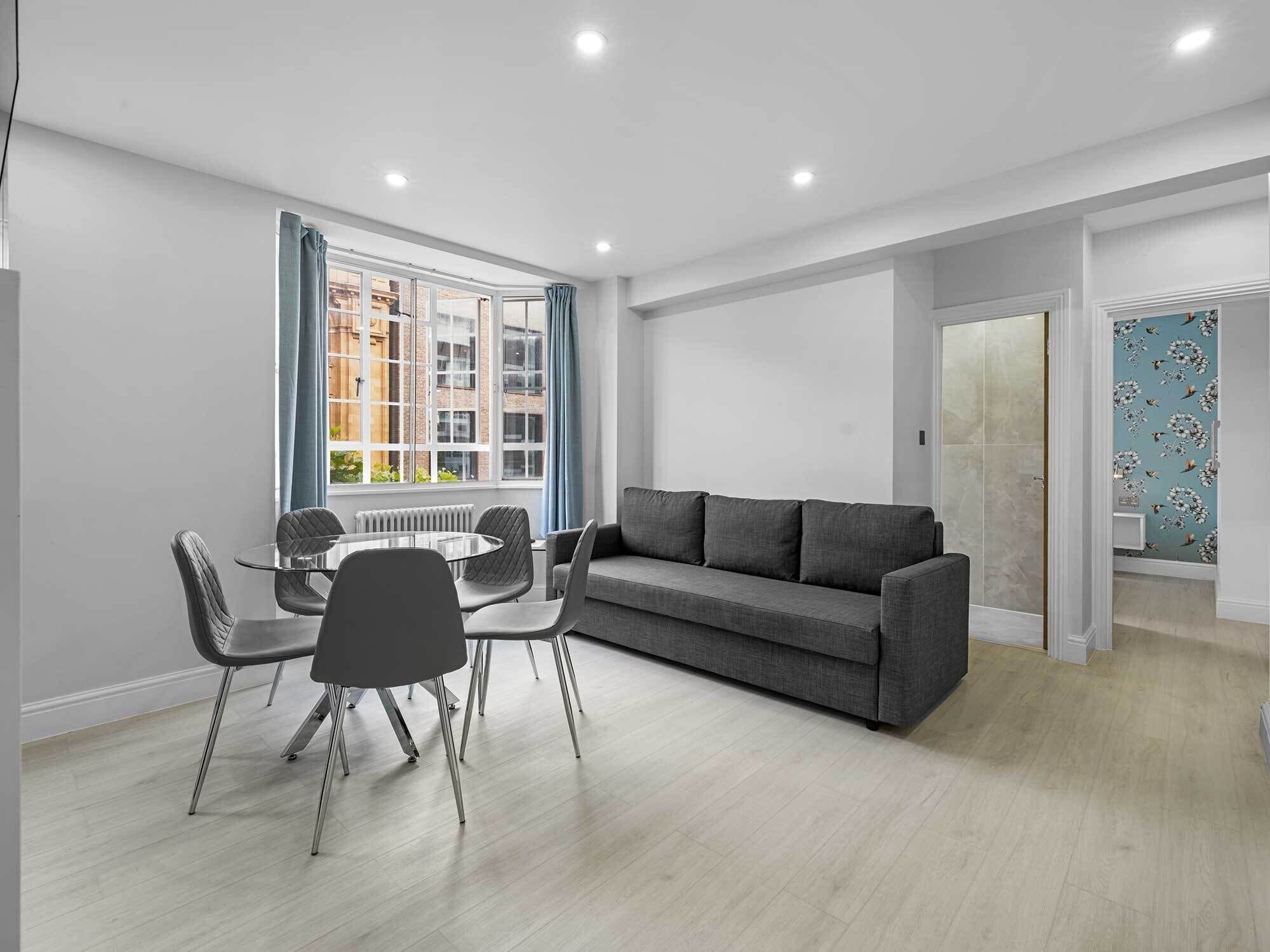 A modern living room with a grey sofa, a glass dining table with chairs, and a large window with curtains.