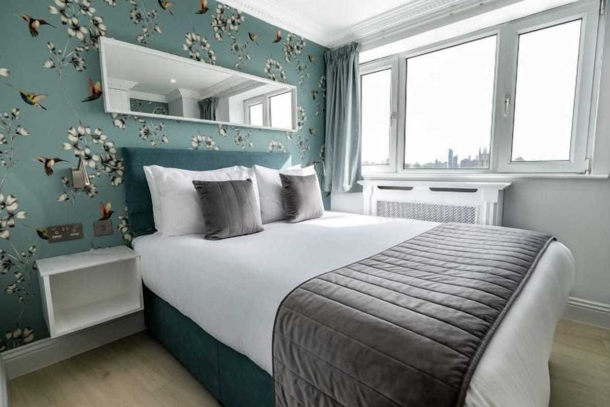 Un dormitorio con cama, mesita de noche, cómoda, espejo y ventana con vista a la ciudad.