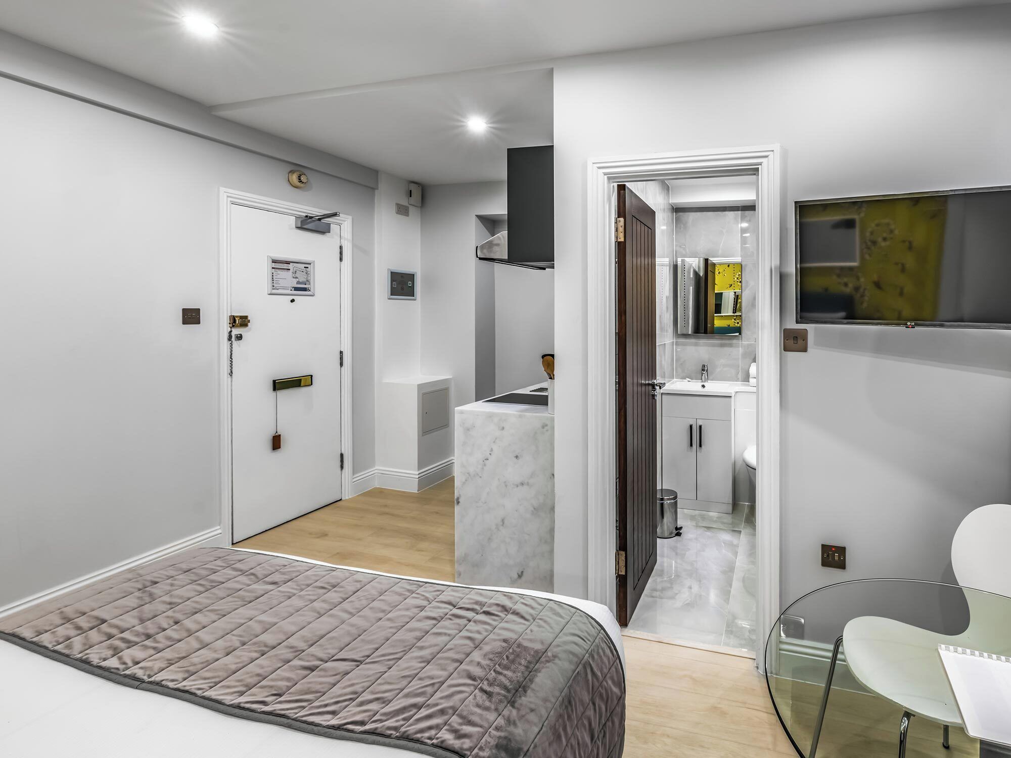 Un dormitorio moderno con una cama, una mesa de vidrio y un baño visible a través de una puerta abierta.