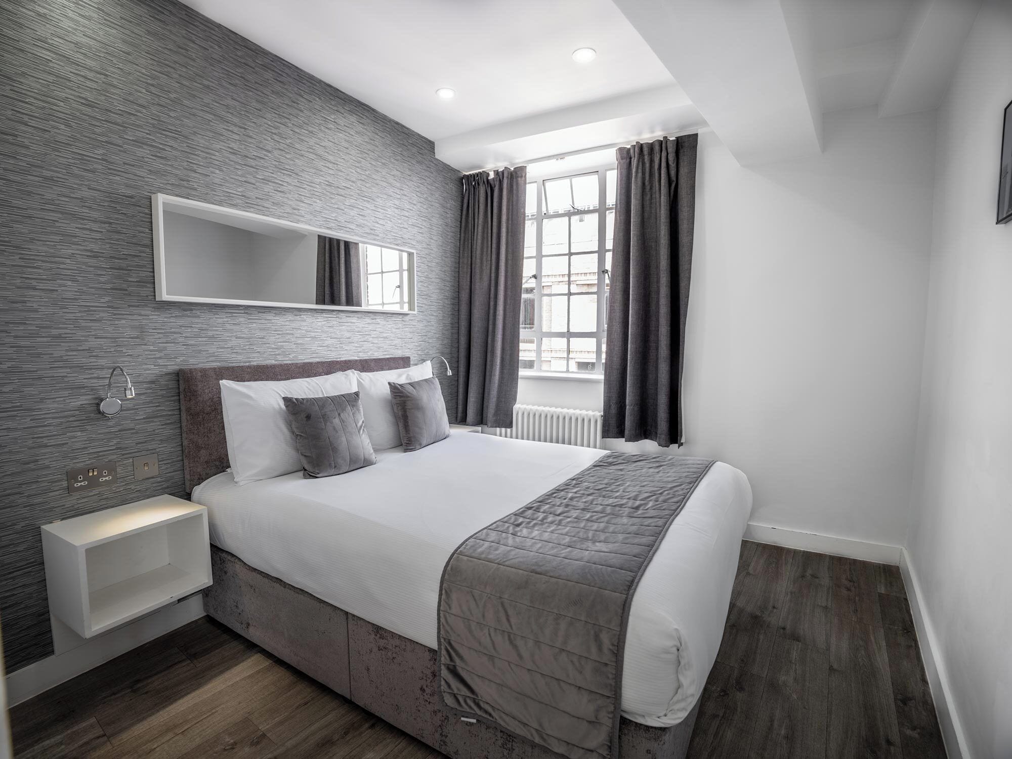 A modern bedroom with a large bed, a bedside table, a wall-mounted light, and a window with curtains.