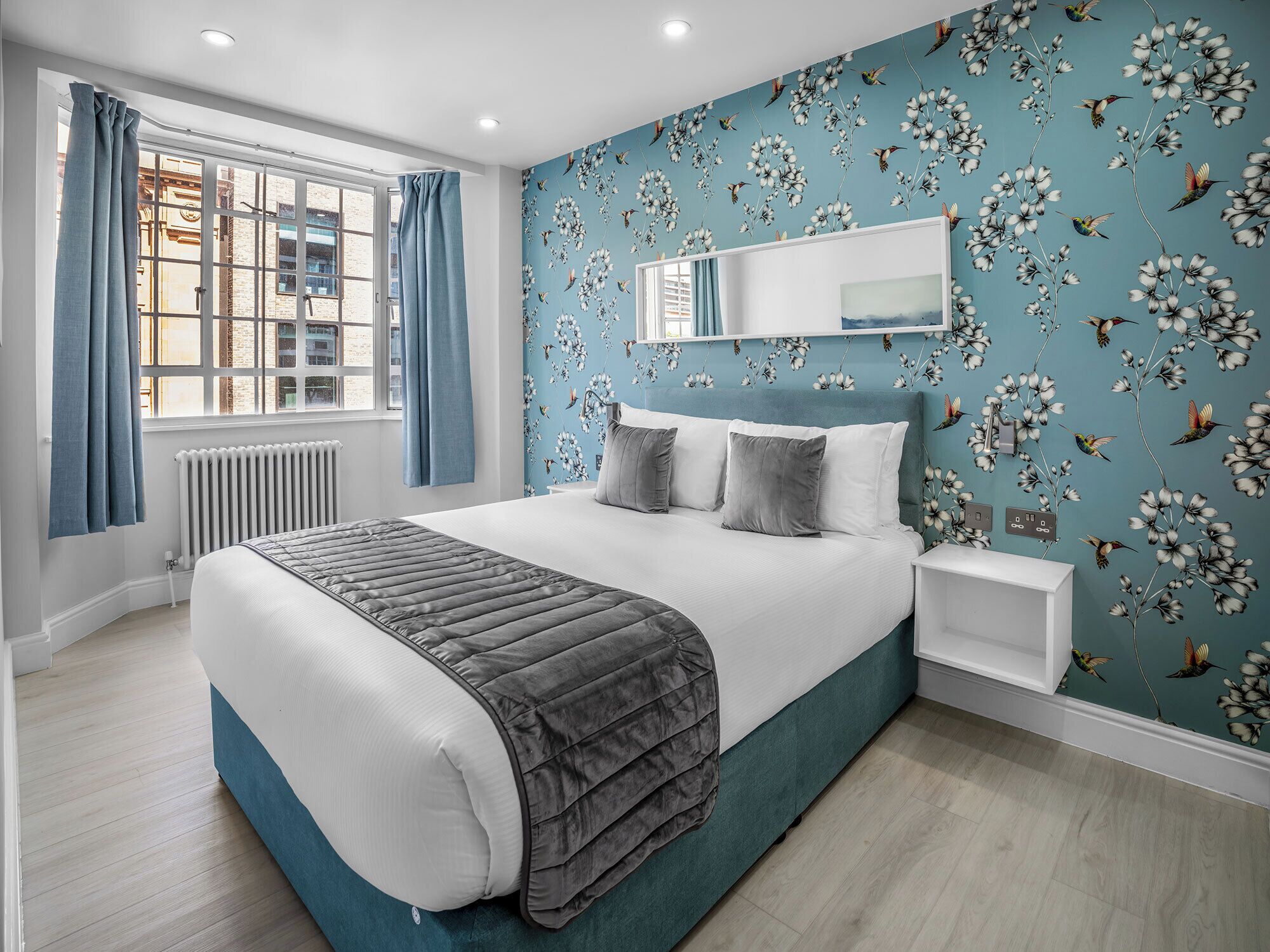 Un dormitorio moderno con una cama grande, una ventana con cortinas, una pared con papel tapiz floral y una mesita de noche.