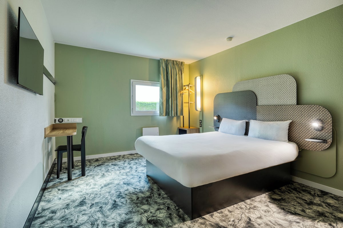 Une chambre d’hôtel moderne avec un grand lit, un bureau avec une chaise, une fenêtre avec des rideaux et une lampe fixée au mur.