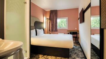 A hotel room with a bed, a desk, a chair, a mirror, and a bathroom with a sink and shower.
