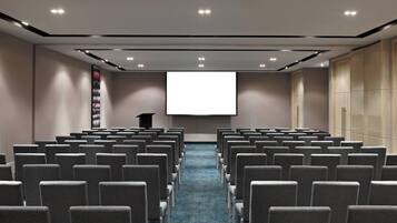 Sala de conferência com fileiras de cadeiras voltadas para uma tela grande, um pódio e piso coberto de carpete.