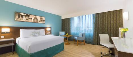 A hotel room with a bed, desk, two chairs, a small table, a painting, and a large window with curtains.