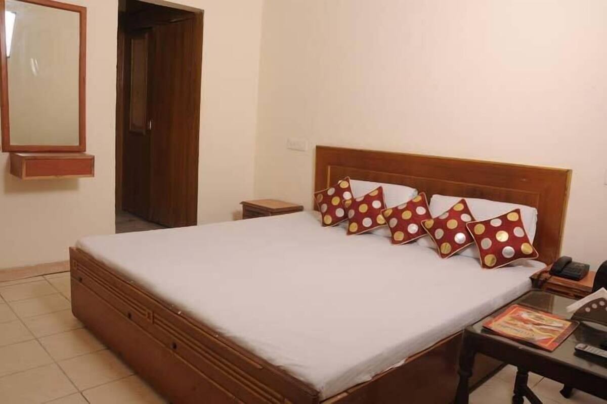 Cama bem arrumada com cabeceira de madeira e quatro almofadas com bolinhas. Uma pequena mesa de cabeceira com abajur e um livro também estão visíveis.