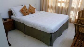 Un lit simple avec un matelas blanc et deux coussins décoratifs, une table de chevet en bois avec une télécommande, une chaise en osier et une fenêtre avec des rideaux.