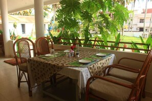 A dining area with a table set for four, surrounded by wicker chairs, with a view of a garden and buildings in the background.