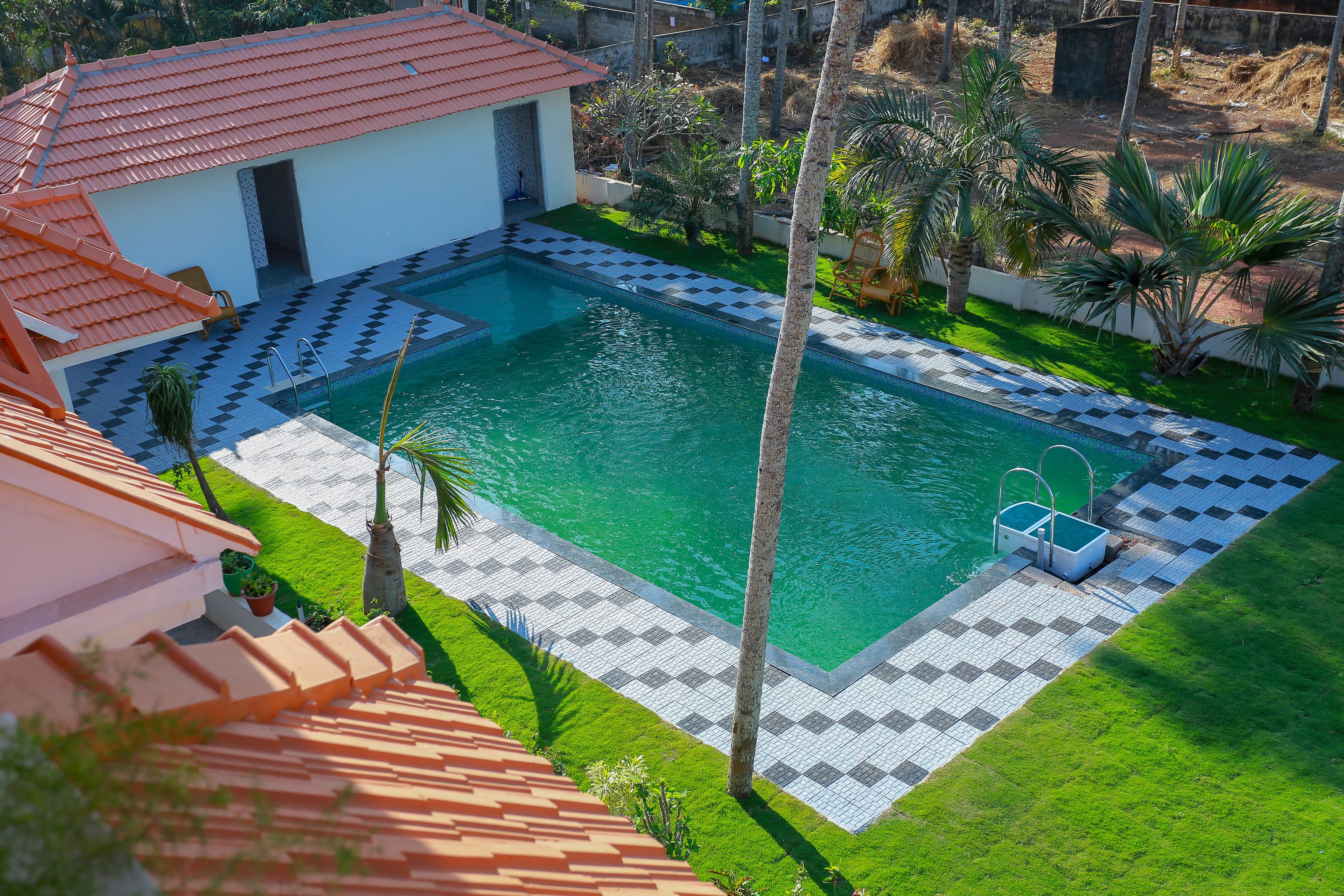Ein Pool mit Schachbrettmuster, ein kleines Haus mit rotem Dach und Palmen.