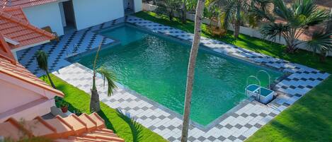 Ein Pool mit Schachbrettmuster, ein kleines Haus mit rotem Dach und Palmen.