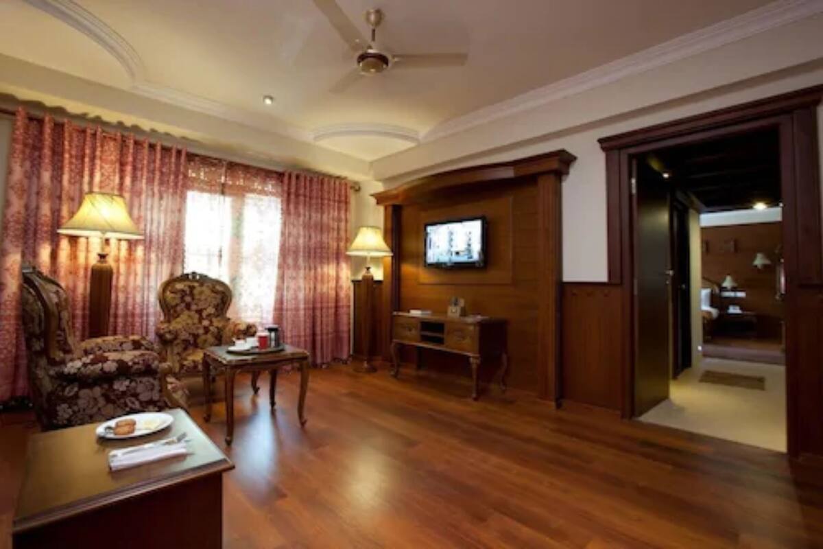 A room with a wooden floor, a sofa, a table with a lamp, a television mounted on the wall, and a door leading to another room.