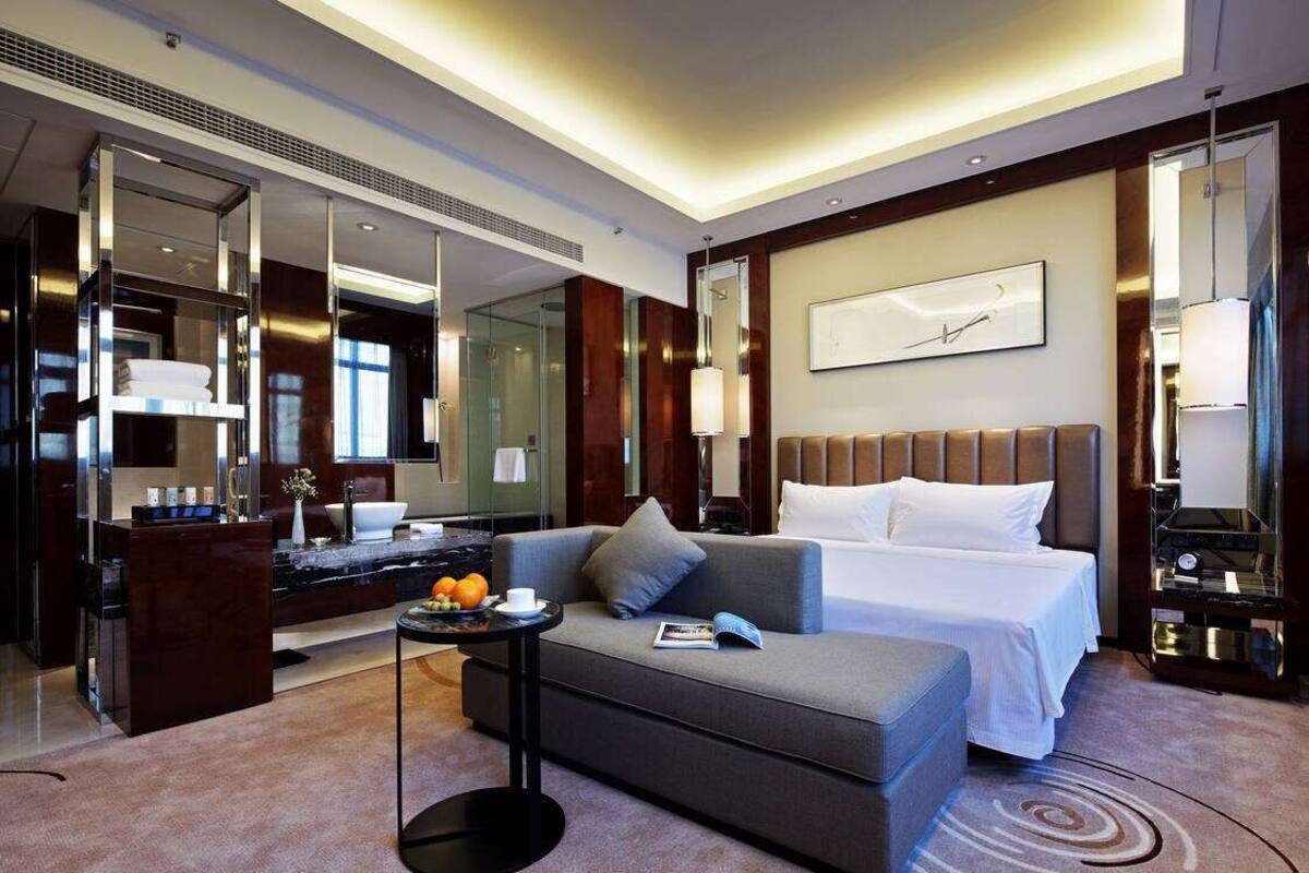 A modern hotel room with a large bed, a sofa, a small table with fruits, a mirror, and a large artwork on the wall.