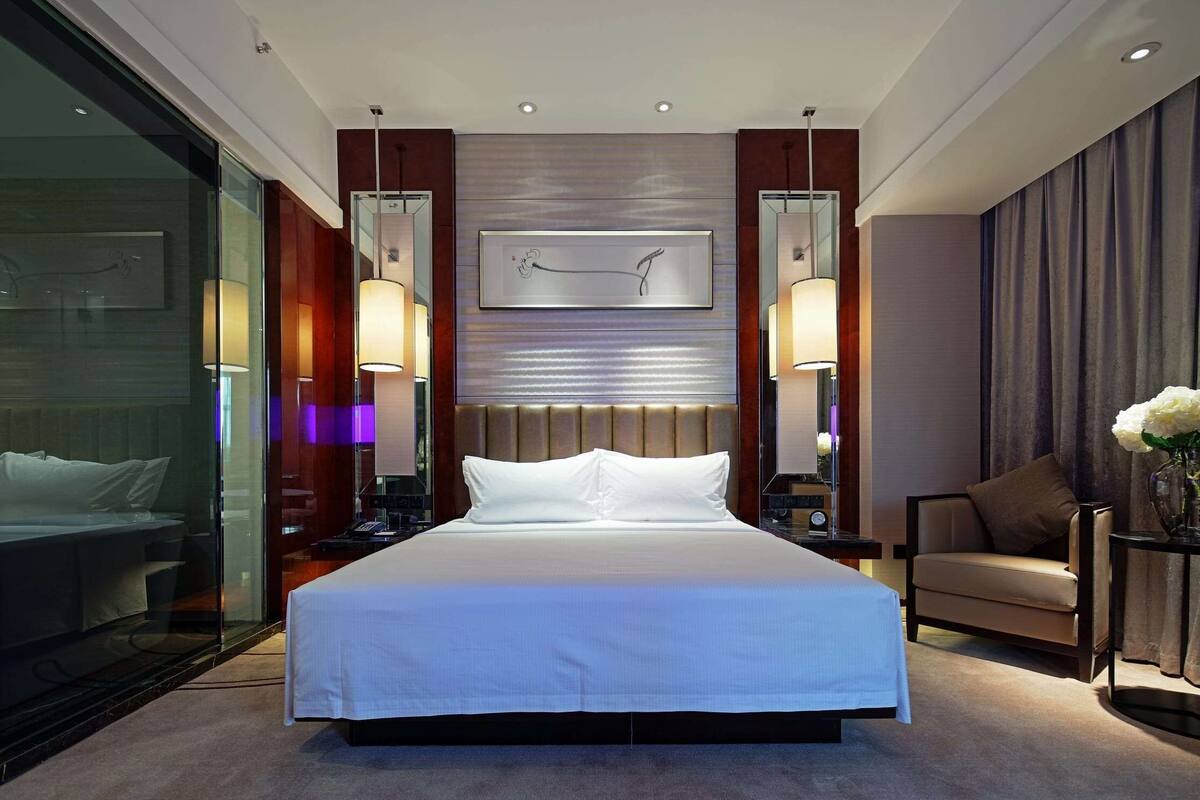 A modern hotel room with a large bed, bedside lamps, a nightstand, a chair, and a wall-mounted artwork.