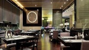 Interior de un restaurante moderno con suelo de madera, mesas y sillas de tonos oscuros, y una obra de arte central en la pared.