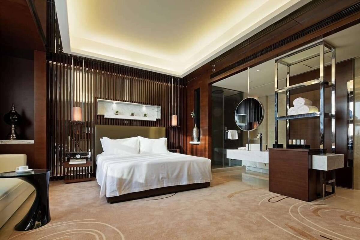 A modern hotel room with a large bed, a desk, a vanity, and a glass shower area.
