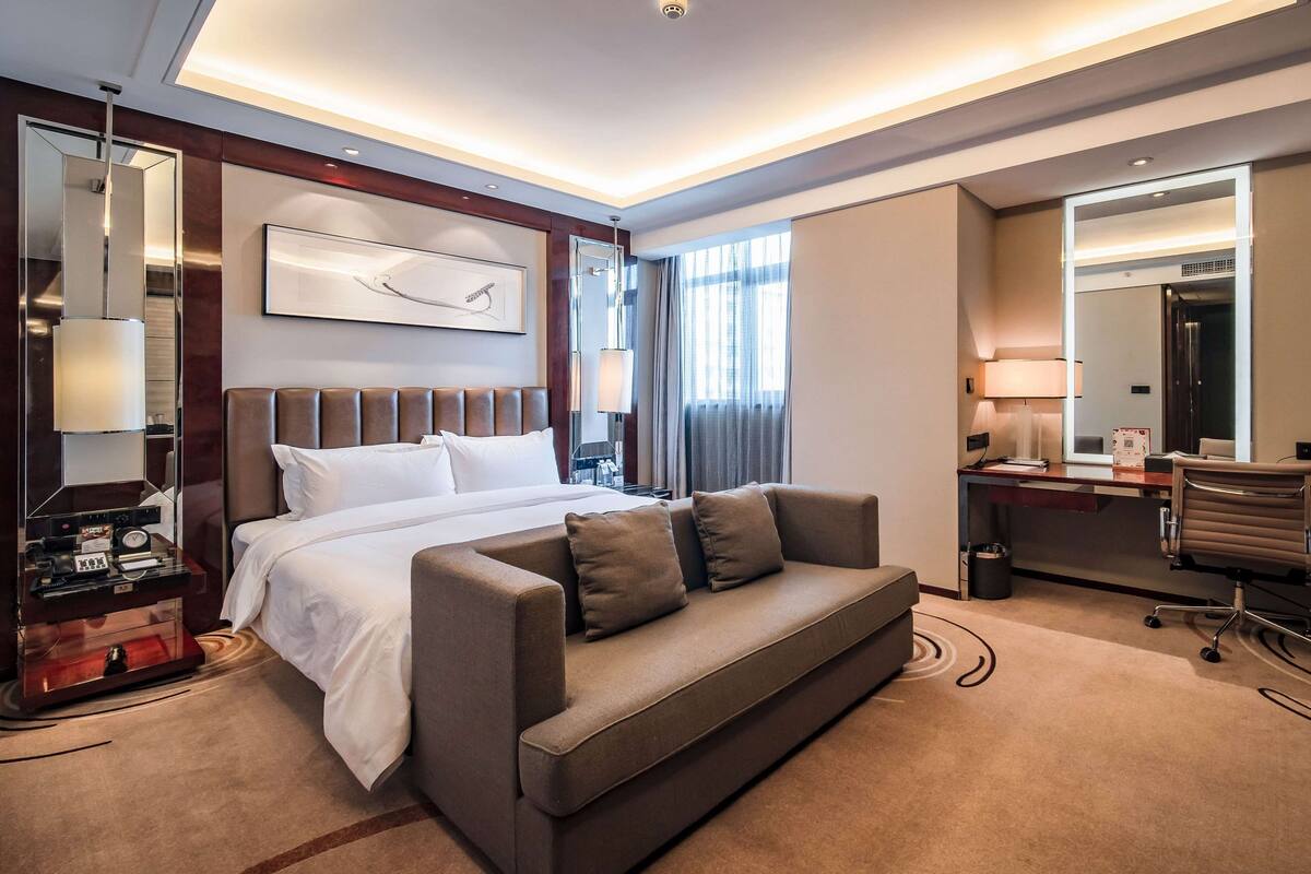A modern hotel room with a large bed, a sofa, a desk, and a mirror.