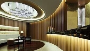 Un moderno vestíbulo de hotel con un mostrador de recepción curvo, una zona de estar con sofá y sillones, y una pared decorativa con paneles verticales.