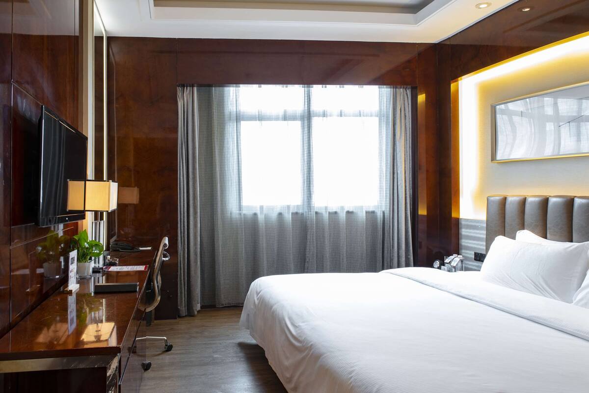 A hotel room with a large bed, a desk with a lamp, a TV, and a window with curtains.