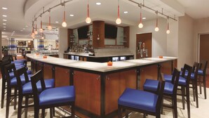 A bar area with a curved counter, multiple hanging light fixtures, and several blue cushioned chairs. - Hilton Garden Inn Falls Church (Falls Church)