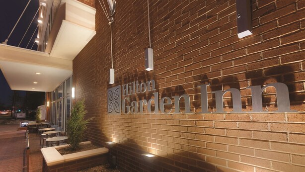 The exterior of a Hilton Garden Inn with a brick facade, outdoor seating area, and illuminated signage. - Hilton Garden Inn Falls Church (Falls Church)
