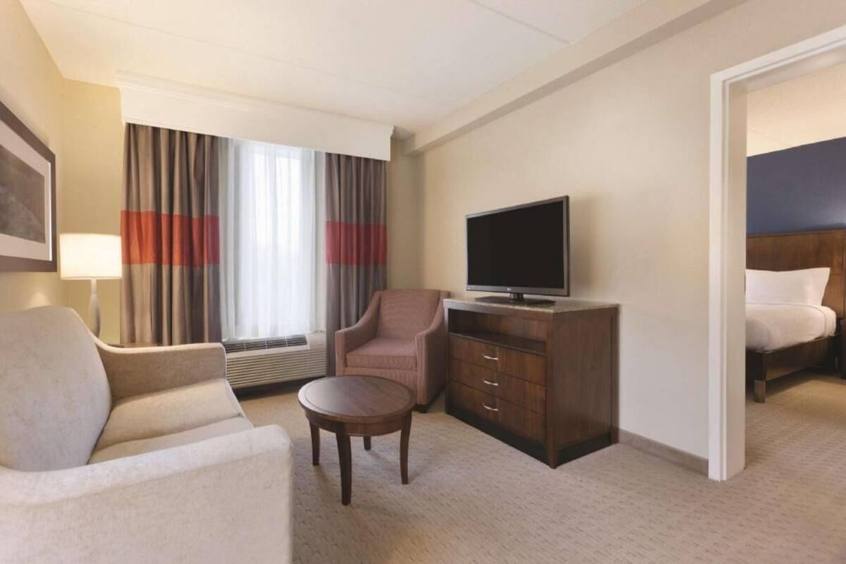 A hotel room with a bed, a TV on a wooden cabinet, a sofa, a chair, a small round table, and a window with curtains.
