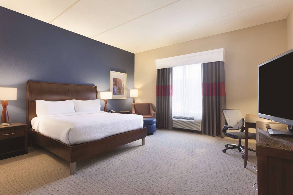 A hotel room with a large bed, a desk with a computer, a chair, a TV, and a window with curtains.