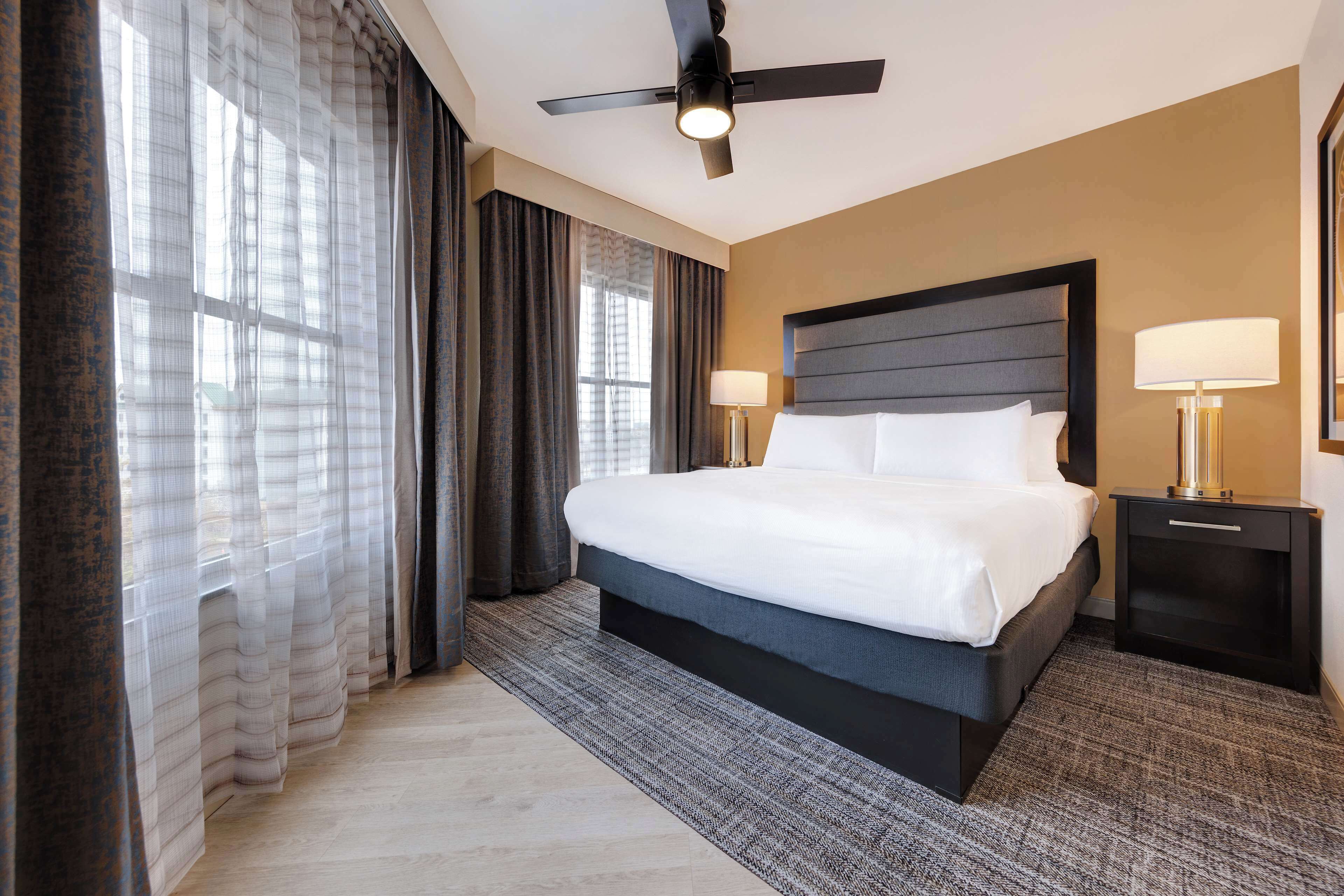 Habitación de hotel con una cama grande, un ventilador de techo, dos lámparas de noche y vistas al exterior a través de cortinas translúcidas.