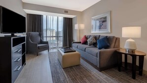 A modern hotel room with a flat-screen TV, a grey sofa, a grey armchair, a small ottoman, a side table with a lamp, and a large window with curtains. - Homewood Suites by Hilton Nashville Vanderbilt, TN (Nashville)