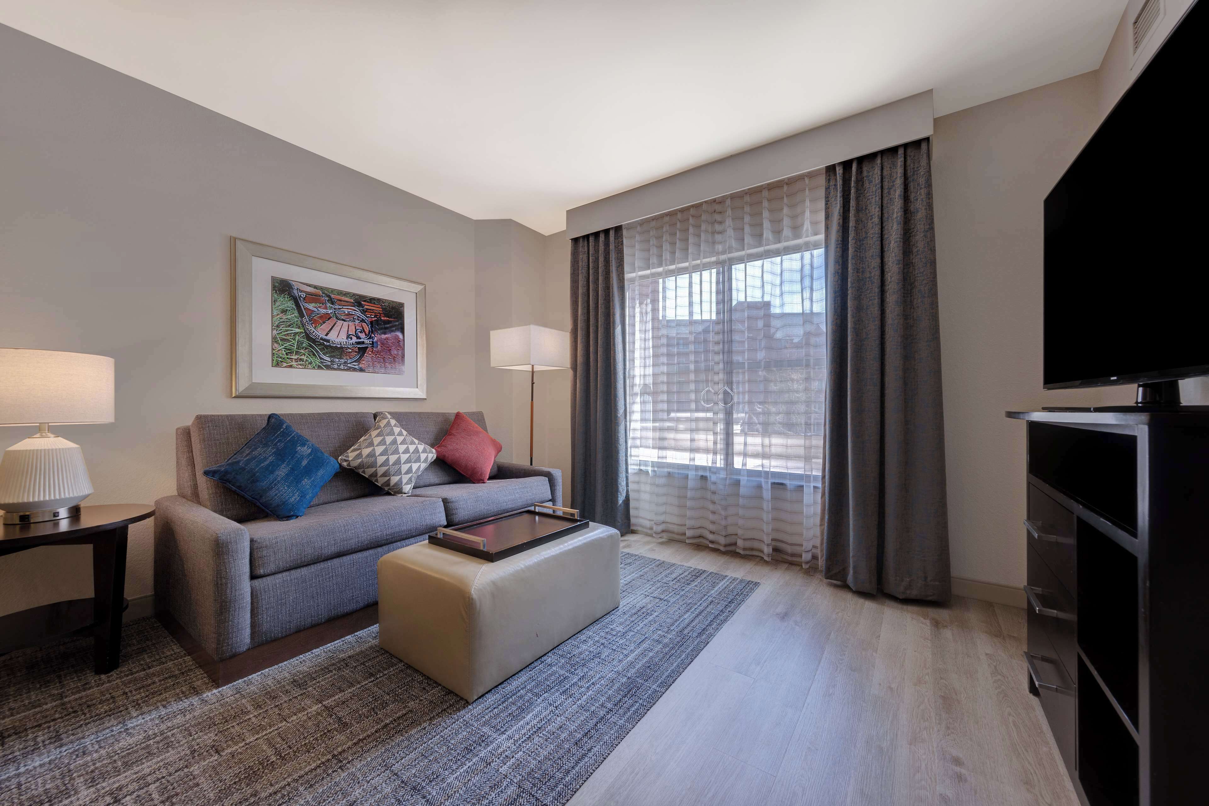 Quarto de hotel moderno com sofá cinza, TV de tela plana, obra de arte emoldurada, luminária e uma janela grande com cortinas.