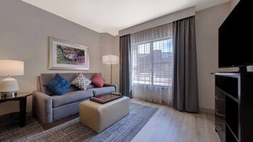 Quarto de hotel moderno com sofá cinza, TV de tela plana, obra de arte emoldurada, luminária e uma janela grande com cortinas.