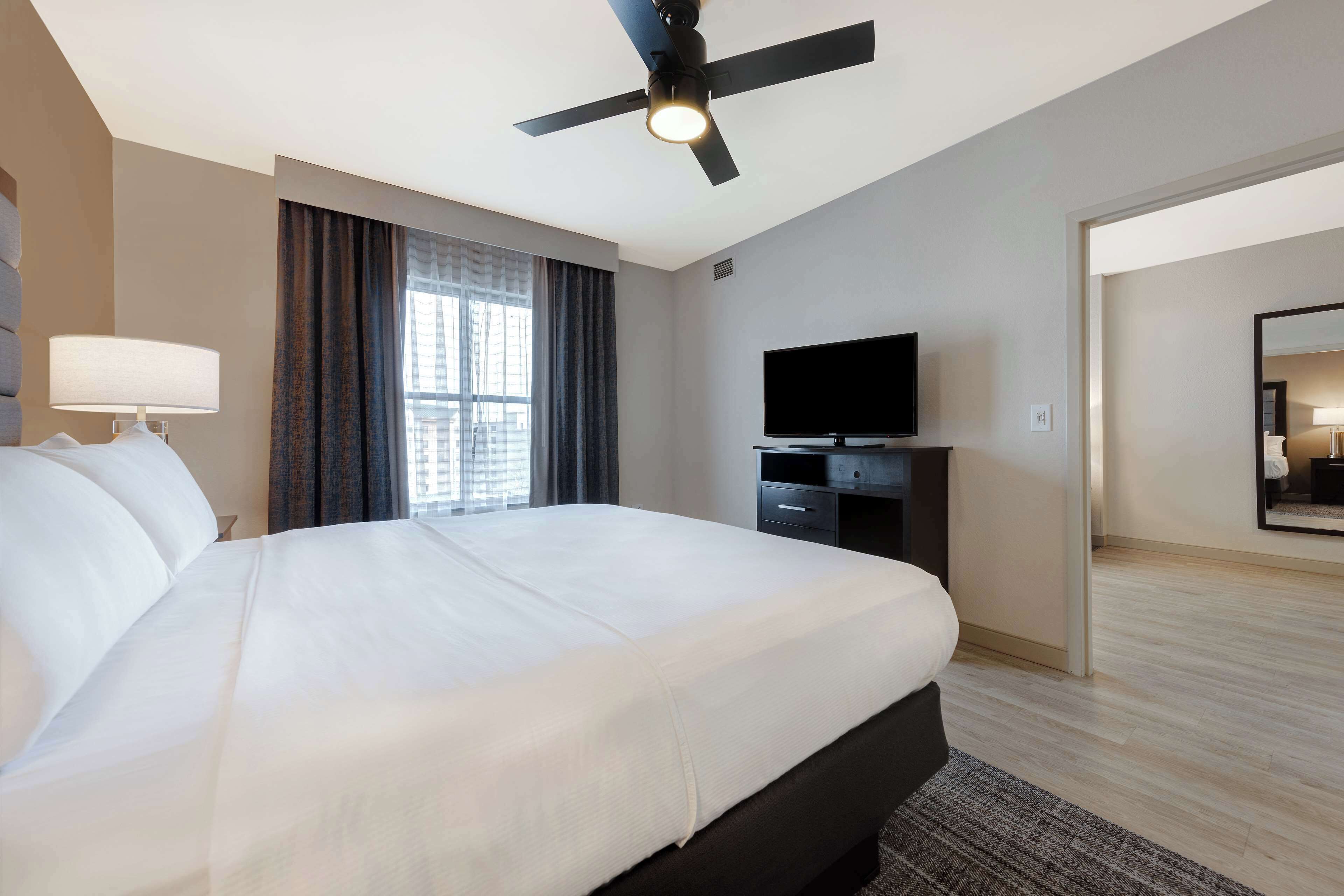 A hotel room with a large bed, a ceiling fan, a flat-screen TV, and a window with curtains.