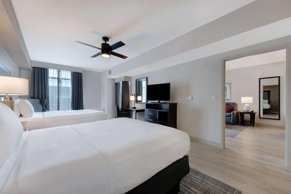 A hotel room with two beds, a ceiling fan, a TV, a desk, and a mirror.