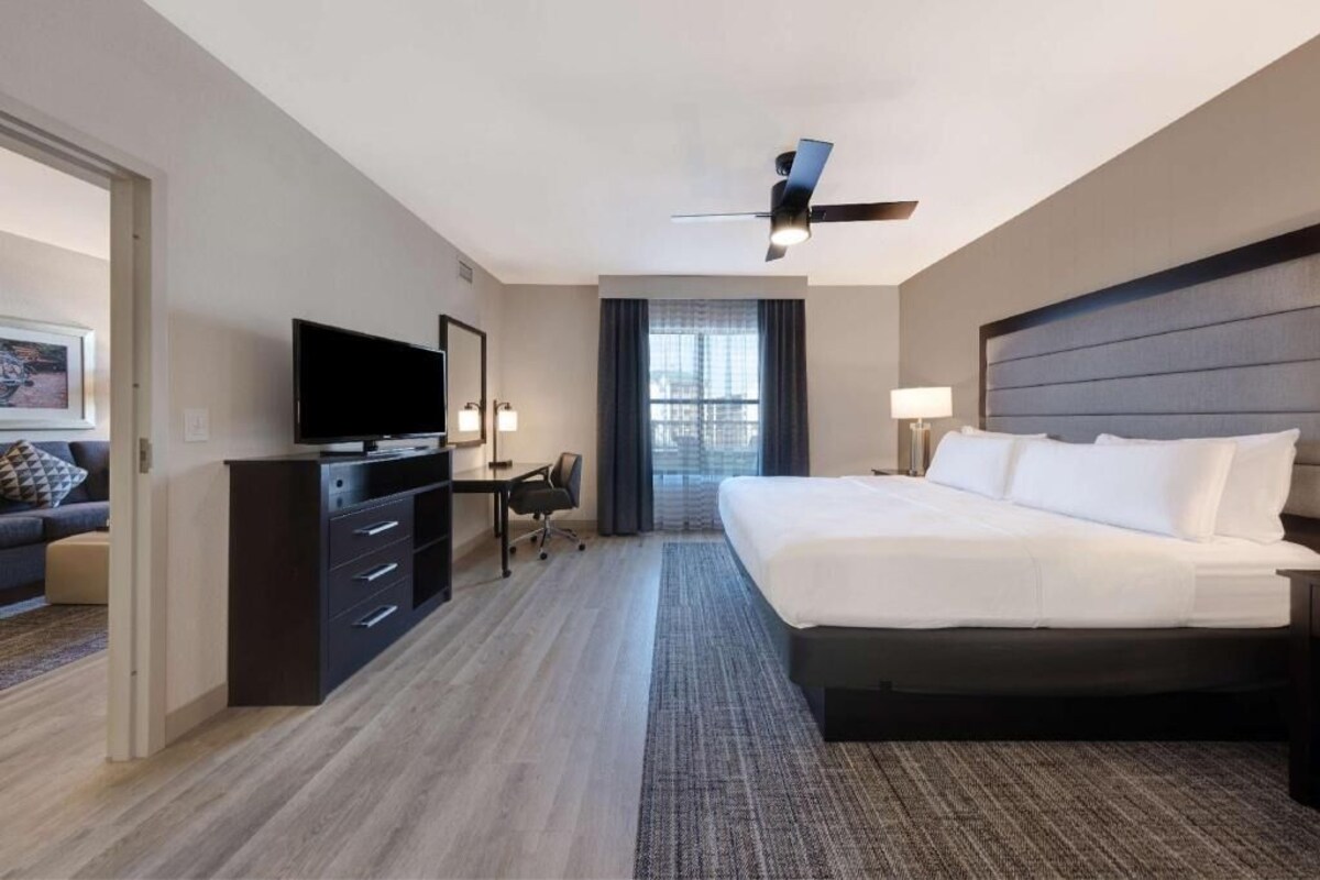 A hotel room with a bed, a desk with a TV, a ceiling fan, and a window with curtains.