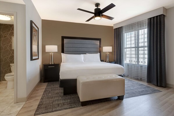A modern hotel room with a large bed, a ceiling fan, a nightstand, a bedside table with lamps, a window with curtains, and a bathroom with a toilet and a sink.