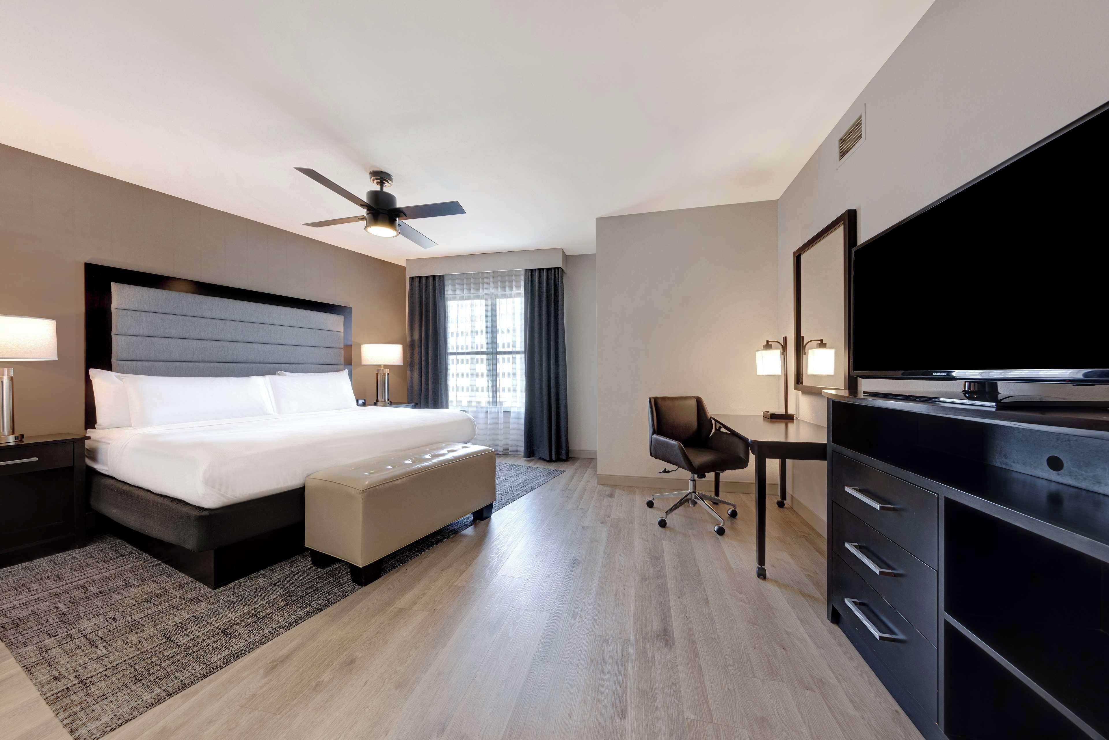 A hotel room with a bed, a desk, a chair, a television, and a ceiling fan.