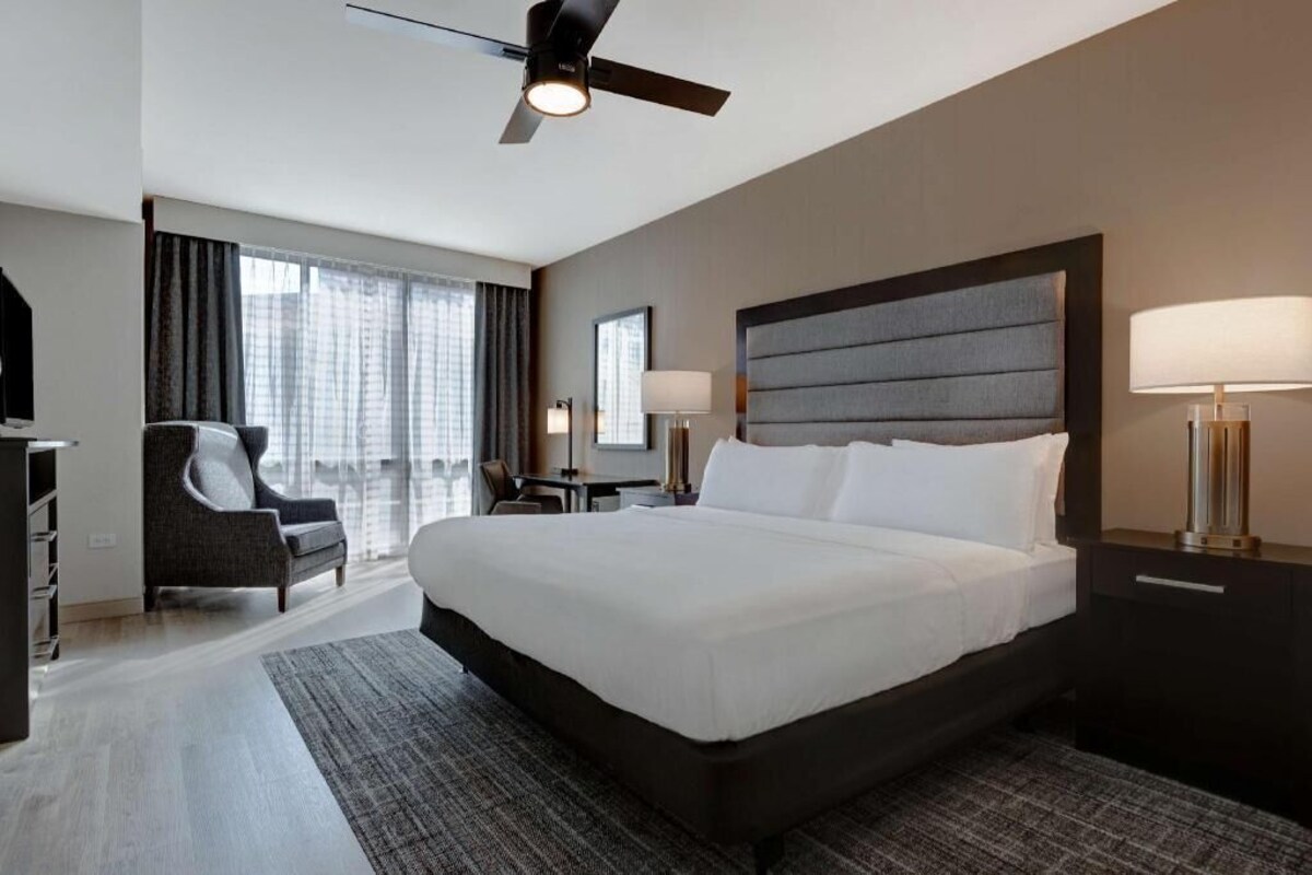 A hotel room with a large bed, a desk, a chair, a ceiling fan, and a view of the city.