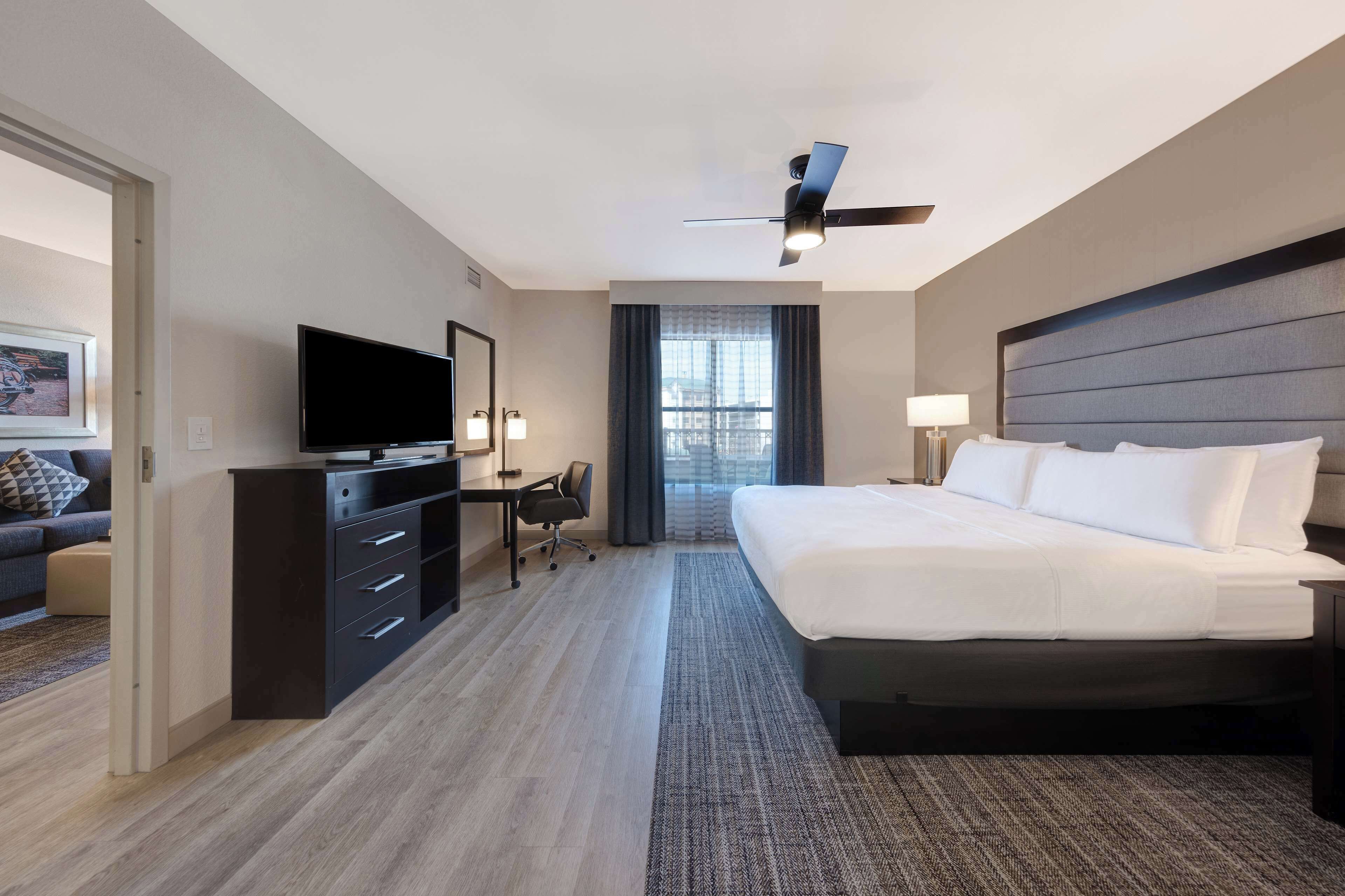 A hotel room with a bed, a desk with a TV, a ceiling fan, and a window with curtains.