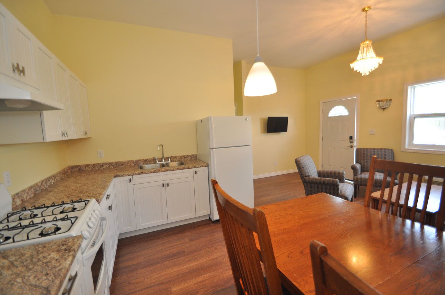 A compact kitchen with white cabinets, a gas stove, and a refrigerator. There is a dining table with chairs and a chandelier above.