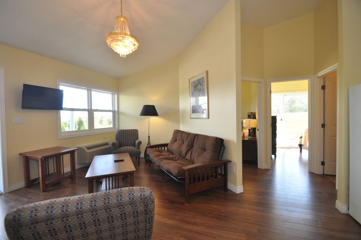 A spacious living room with hardwood floors, a brown leather sofa, a patterned armchair, and a wooden coffee table. A chandelier hangs from the ceiling, and a flat-screen TV is mounted on the wall.